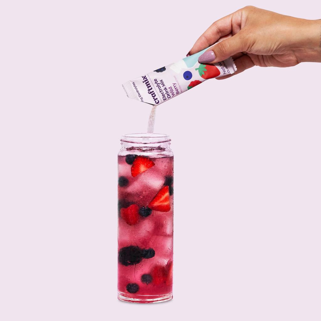 Hand pouring Craftmix Wild Berry Electrolyte Drink Mix into a glass of ice water with fresh strawberries, blueberries, and blackberries.