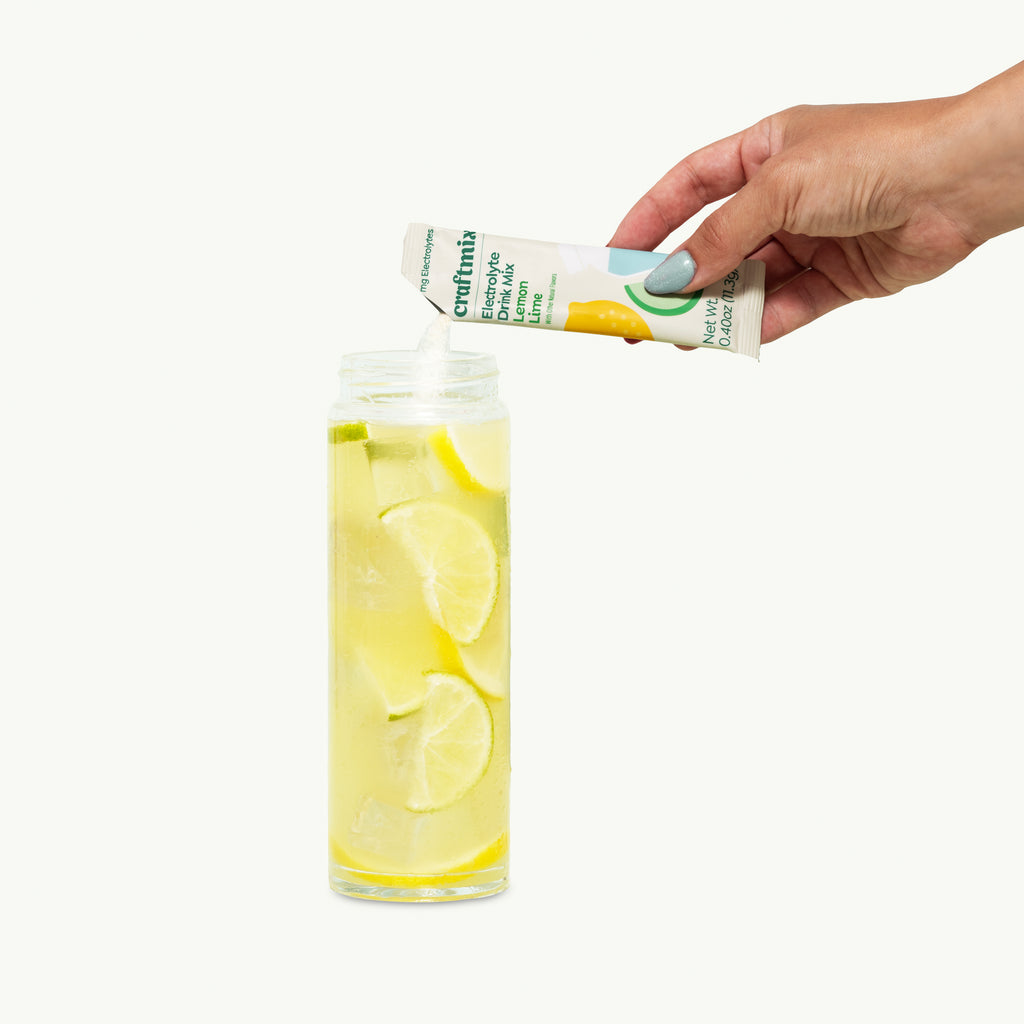 Hand pouring Craftmix Lemon Lime Electrolyte Drink Mix into a glass of ice water with fresh lemon and lime slices.
