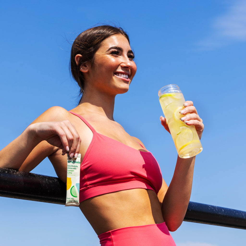 Woman holding a Craftmix Lemon Lime Electrolyte Drink Mix stick pack and a glass of lemon lime electrolyte water outdoors.