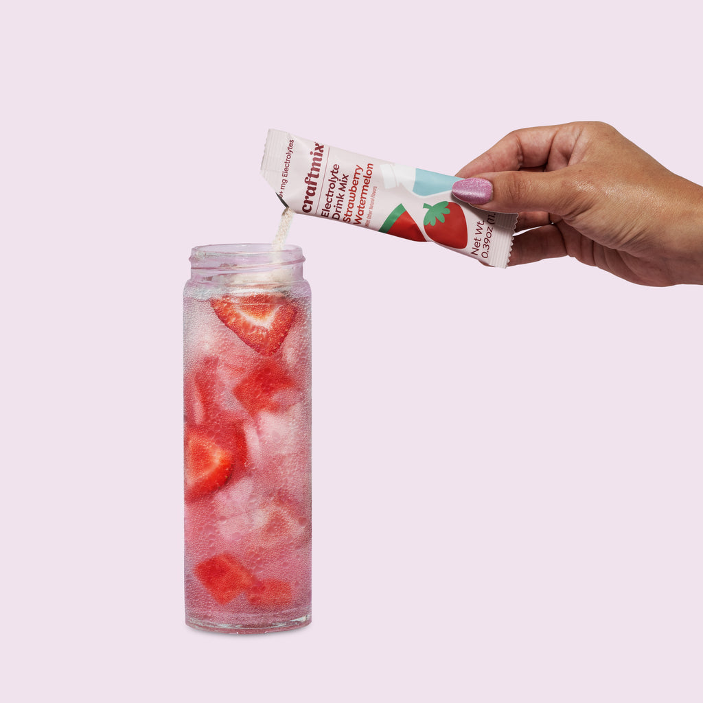 Hand pouring Craftmix Strawberry Watermelon Electrolyte Drink Mix into a glass of ice water with fresh strawberry slices.